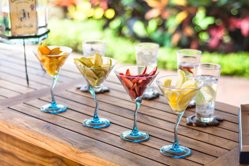 Alcoholic drinks in a variety of clear glasses with fruit in them, sitting on a wooden table.